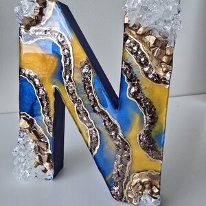 Puede incluir: Una letra "N" decorativa con un dise&ntilde;o de geoda azul, amarillo y plateado. La letra est&aacute; adornada con detalles dorados y adornos transparentes similares a cristales. La letra mide aproximadamente 20 cm de alto.