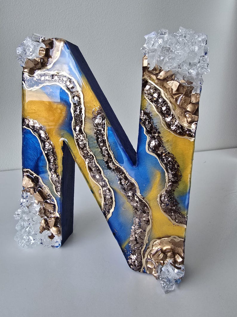 Puede incluir: Una letra "N" decorativa con un dise&ntilde;o de geoda azul, amarillo y plateado. La letra est&aacute; adornada con detalles dorados y adornos transparentes similares a cristales. La letra mide aproximadamente 20 cm de alto.