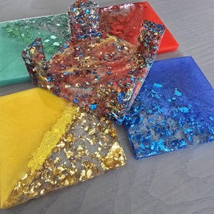 Pode incluir: Um conjunto de bases para copos de resina colorida com flocos met&aacute;licos embutidos. As bases s&atilde;o de v&aacute;rias formas e cores, incluindo vermelho, azul, amarelo e verde. Inclui um suporte decorativo central.