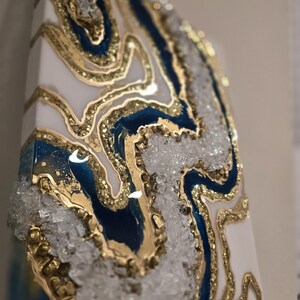 Op de afbeelding: Abstract kunstwerk met een geode-ontwerp. Het kunstwerk heeft een witte basis met blauwe en gouden accenten en heldere kristallen versieringen. De gouden elementen cre&euml;ren een gelaagd effect, waardoor diepte en textuur aan het stuk worden toegevoegd.