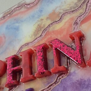 Puede incluir: Primer plano de una pieza de arte decorativa de resina con la palabra "FIN" en letras rojas llenas de purpurina. El fondo presenta un dise&ntilde;o colorido y veteado con detalles de purpurina rosa y los n&uacute;meros "2025" en morado.