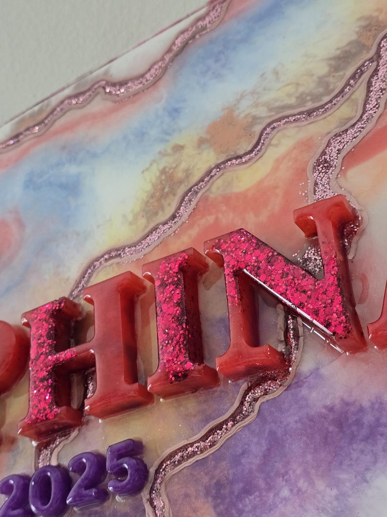 Puede incluir: Primer plano de una pieza de arte decorativa de resina con la palabra "FIN" en letras rojas llenas de purpurina. El fondo presenta un dise&ntilde;o colorido y veteado con detalles de purpurina rosa y los n&uacute;meros "2025" en morado.