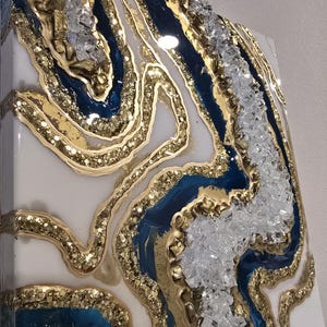 Op de afbeelding: Abstract kunstwerk van hars met een geode-ontwerp. Het kunstwerk heeft lagen van wit, diepblauw en goudkleurig glitterhars, met heldere kristalaccenten. Het heeft een glanzende afwerking en een getextureerd uiterlijk.