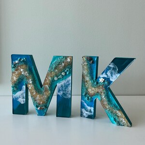 Może przedstawiać: Dwie litery z żywicy, "M" i "K", w odcieniach niebieskiego i turkusowego, z motywem plażowym. Litery wypełnione są piaskiem, muszlami i przezroczystą żywicą, tworząc efekt 3D. Litery mają około 15 cm wysokości.