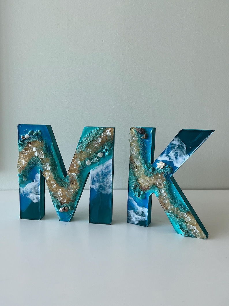 Może przedstawiać: Dwie litery z żywicy, "M" i "K", w odcieniach niebieskiego i turkusowego, z motywem plażowym. Litery wypełnione są piaskiem, muszlami i przezroczystą żywicą, tworząc efekt 3D. Litery mają około 15 cm wysokości.