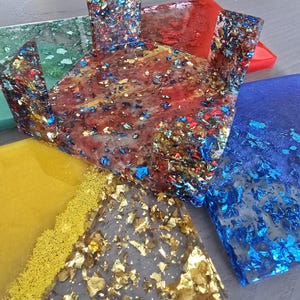 Pode incluir: Uma cole&ccedil;&atilde;o de pe&ccedil;as de arte em resina coloridas. As pe&ccedil;as t&ecirc;m v&aacute;rias formas, incluindo uma caixa e azulejos retangulares. A resina &eacute; infundida com flocos met&aacute;licos dourados, azuis e vermelhos. As cores incluem vermelho, azul, verde, amarelo e transparente.