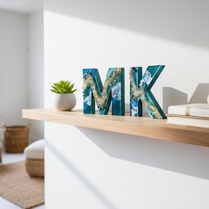 Może przedstawiać: Ozdobne drewniane litery z napisem "MK" z niebieskim i złotym wzorem z żywicy, wystawione na drewnianej p&oacute;łce. Mały sukulent w białej doniczce, muszla i książki r&oacute;wnież znajdują się na p&oacute;łce.