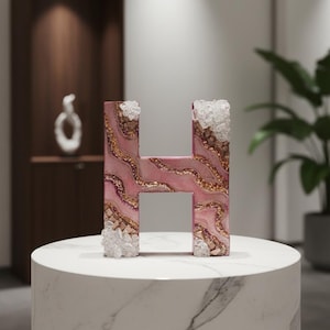 May include: A decorative letter "H" with a pink, gold, and white marbled design. The letter is embellished with clear crystal-like accents on the top and bottom. It is displayed on a white marble pedestal.