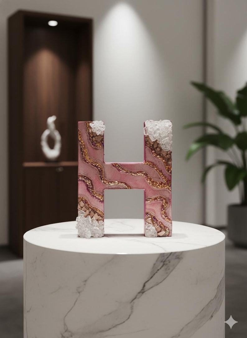 May include: A decorative letter "H" with a pink, gold, and white marbled design. The letter is embellished with clear crystal-like accents on the top and bottom. It is displayed on a white marble pedestal.