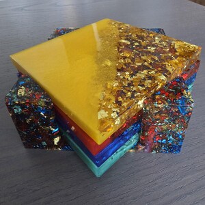 Pode incluir: Um conjunto de bases de resina coloridas. A base superior &eacute; amarela com detalhes em folha de ouro. Abaixo, camadas vermelhas, azuis e turquesas. As laterais s&atilde;o feitas de resina com flocos coloridos.