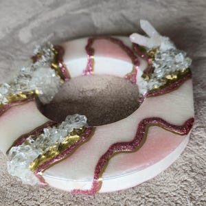 May include: A decorative, circular object with a white base, accented with pink and gold glitter details. Clear crystal-like embellishments are clustered around the edges, creating a unique, artistic design.