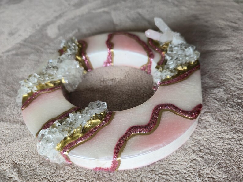 May include: A decorative, circular object with a white base, accented with pink and gold glitter details. Clear crystal-like embellishments are clustered around the edges, creating a unique, artistic design.