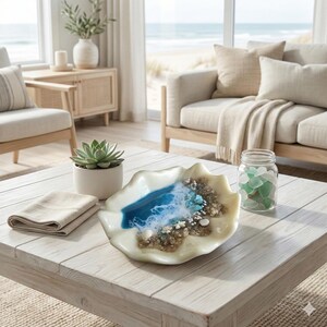 May include: A decorative seashell-shaped dish with a blue and white resin ocean scene, featuring sand, shells, and turquoise accents. A small succulent in a white pot, a folded beige napkin, and a jar of sea glass are also on the white wooden table.