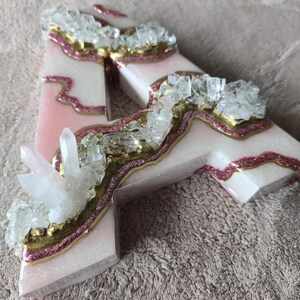 May include: A decorative letter "A" with a pink and white marbled design, accented with gold glitter and clear crystal clusters. The letter is a unique, handcrafted piece.