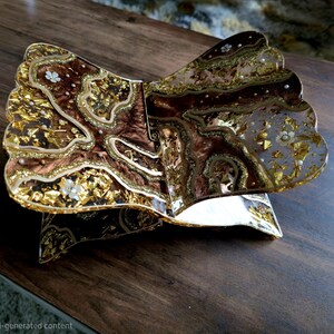 May include: An open, decorative book with a gold and brown design. The book has a layered, textured appearance with gold leaf accents and small white floral embellishments. The edges of the pages have a scalloped design. The book is resting on a dark wooden surface.