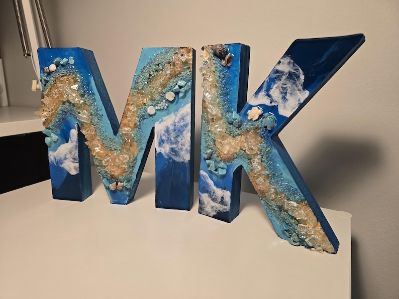 Może przedstawiać: Dekoracyjne drewniane litery składające się na napis "MK", pomalowane na niebiesko i ozdobione żywicą, kryształami i muszlami. Litery mają motyw nadmorski, z wzorami fal i osadzonymi kamieniami. Unikalny element wystroju domu.