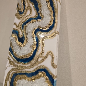 May include: Abstract resin art piece with a geode design. The artwork features a white background with blue and gold accents, and clear crystal-like embellishments. The piece is rectangular and has a textured surface.