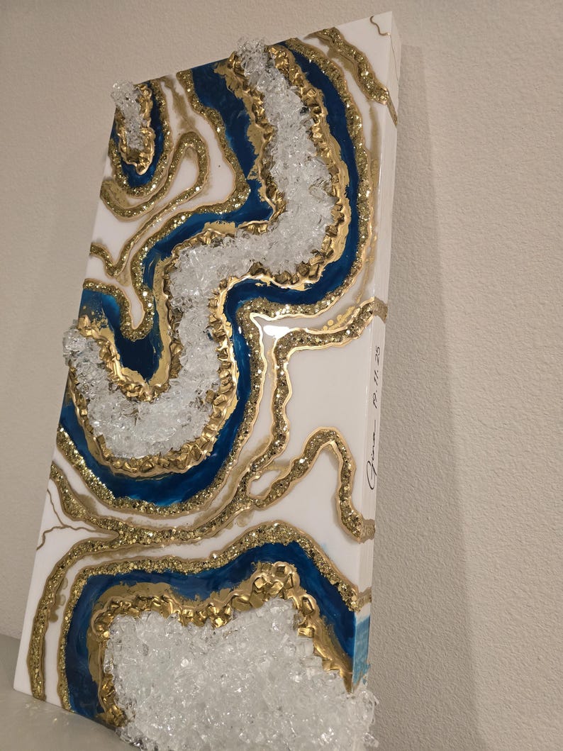 May include: Abstract resin art piece with a geode design. The artwork features a white background with blue and gold accents, and clear crystal-like embellishments. The piece is rectangular and has a textured surface.