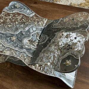 May include: A decorative, open book-shaped tray with a silver and gray marbled design. The tray has a scalloped edge and is embellished with silver leaf and small floral accents. The tray is made of resin.