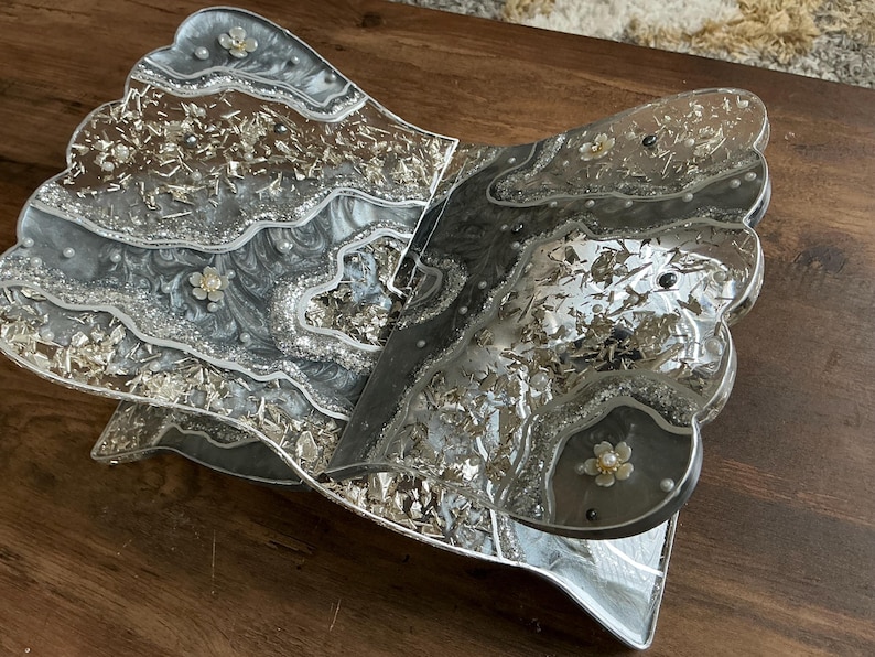 May include: A decorative, open book-shaped tray with a silver and gray marbled design. The tray has a scalloped edge and is embellished with silver leaf and small floral accents. The tray is made of resin.