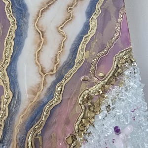 Może przedstawiać: Abstrakcyjne dzieło sztuki z motywem geody. Zawiera warstwy białej, niebieskiej i r&oacute;żowej żywicy, zaakcentowane złotym brokatem i złotymi płatkami. Prawa strona ma przezroczyste i fioletowe kryształy.