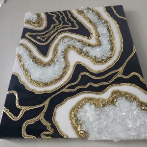 May include: Abstract wall art with a geode design. The artwork features a black, white, and gold colour scheme with clear crystal accents. The piece has a glossy finish and is mounted on a wall.