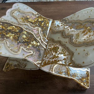 May include: A decorative, bow-shaped resin tray with a layered design. The tray features a creamy white base with gold leaf accents and embedded pearls. The edges are outlined with gold glitter, and the tray has a scalloped edge.
