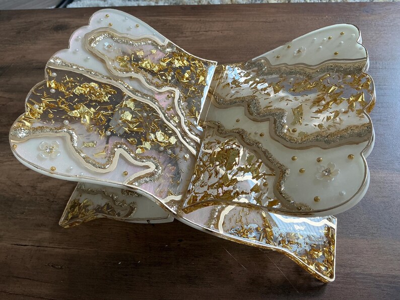 May include: A decorative, bow-shaped resin tray with a layered design. The tray features a creamy white base with gold leaf accents and embedded pearls. The edges are outlined with gold glitter, and the tray has a scalloped edge.