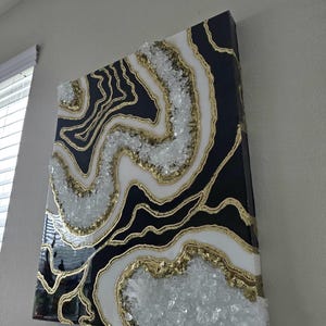 May include: Abstract geode-style wall art in black, white, and gold. The design incorporates crystal-like elements and gold outlines, creating a layered effect. The artwork is mounted on a black canvas, offering a striking contrast. The piece is a decorative item.