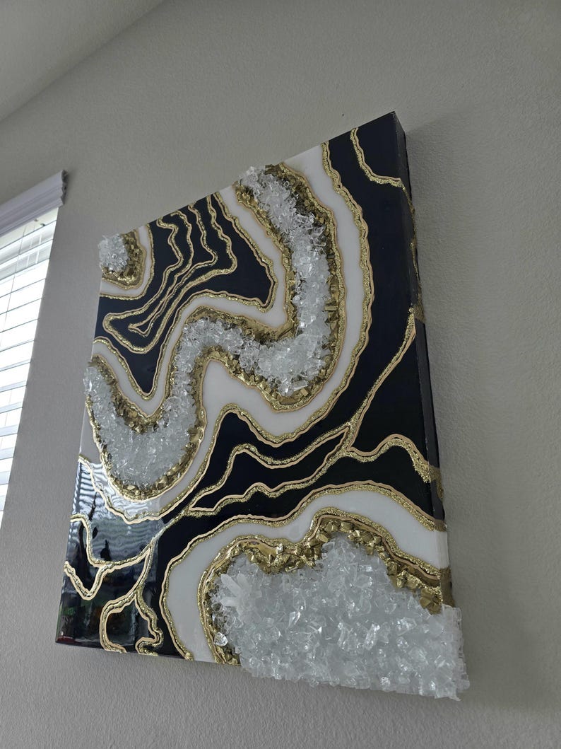 May include: Abstract geode-style wall art in black, white, and gold. The design incorporates crystal-like elements and gold outlines, creating a layered effect. The artwork is mounted on a black canvas, offering a striking contrast. The piece is a decorative item.