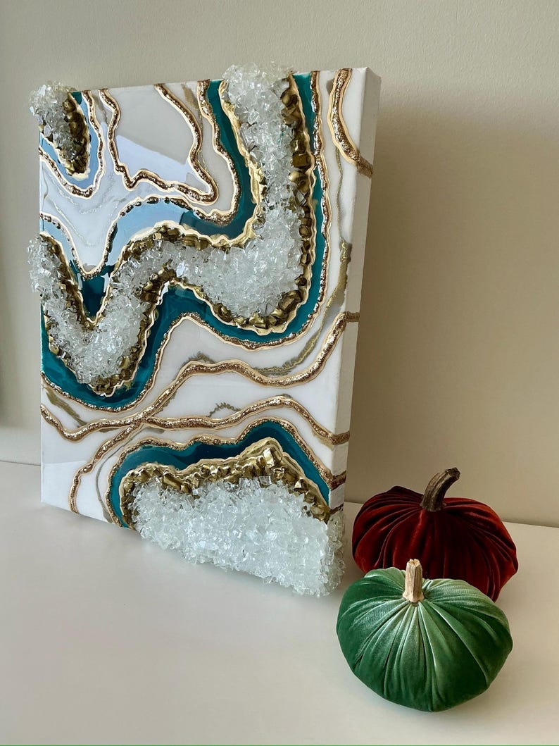 May include: An abstract canvas art piece with a geode design. The artwork features layers of white, teal, gold, and clear crystal accents. Two velvet pumpkins, one red and one green, are in the foreground.