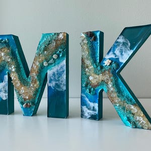 May include: Decorative resin letters spelling "MK". The letters are teal with a beach-themed design, incorporating white, tan, and clear elements to mimic sand, waves, and shells. The letters are displayed on a white surface.