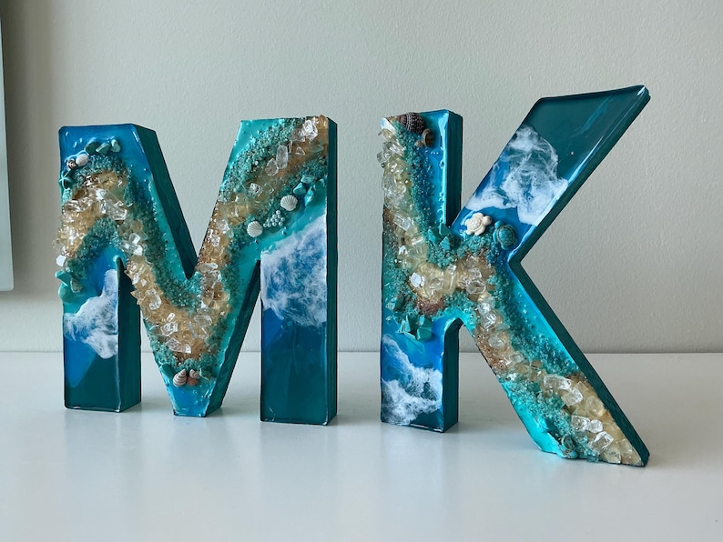 May include: Decorative resin letters spelling "MK". The letters are teal with a beach-themed design, incorporating white, tan, and clear elements to mimic sand, waves, and shells. The letters are displayed on a white surface.