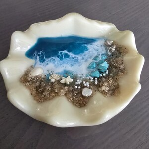 May include: An off-white seashell-shaped dish featuring a miniature beach scene. The scene includes a blue and white ocean, sand, turquoise stones, pearls, and tiny sea creatures. A unique coastal-themed decorative item.