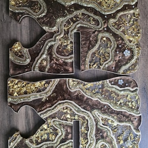 May include: Two decorative resin letters, each with a brown base, gold leaf accents, and glitter details. The letters have a unique, organic shape with a wavy design and small white flower embellishments.
