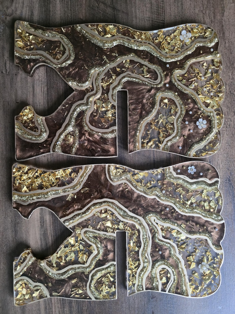 May include: Two decorative resin letters, each with a brown base, gold leaf accents, and glitter details. The letters have a unique, organic shape with a wavy design and small white flower embellishments.
