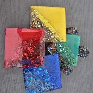 Pode incluir: Arranjo de pe&ccedil;as de arte em resina coloridas. Os quadrados s&atilde;o em tons de amarelo, vermelho, azul e verde, com flocos de ouro e outros metais embutidos. As pe&ccedil;as est&atilde;o dispostas sobre uma superf&iacute;cie cinza.