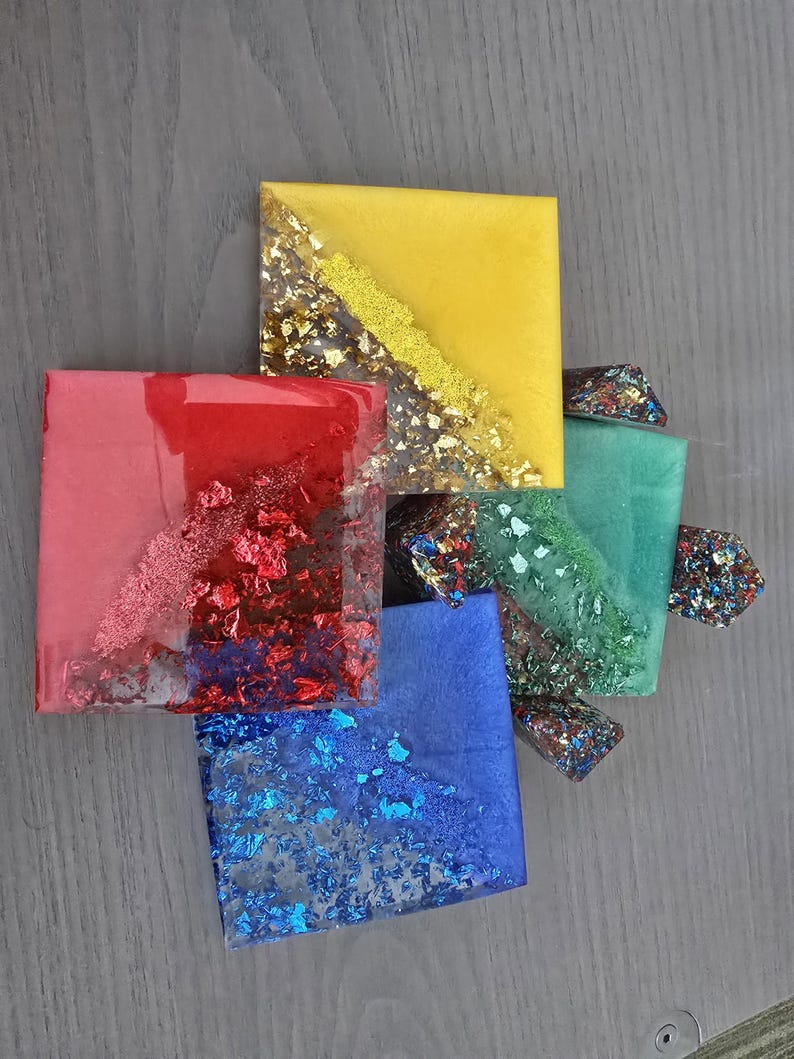 Pode incluir: Arranjo de pe&ccedil;as de arte em resina coloridas. Os quadrados s&atilde;o em tons de amarelo, vermelho, azul e verde, com flocos de ouro e outros metais embutidos. As pe&ccedil;as est&atilde;o dispostas sobre uma superf&iacute;cie cinza.