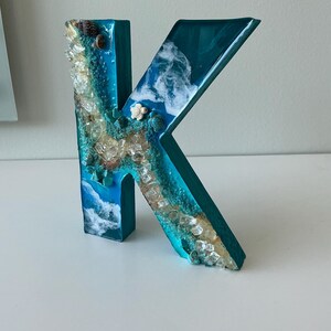 Może przedstawiać: Turkusowa drewniana litera "K" ozdobiona żywicą i elementami morskimi. Litera ma turkusową podstawę z żywicy z zatopionymi muszlami, przezroczystymi kryształami i białymi wzorami fal, tworzącymi estetykę nadmorską. Litera ma około 20 cm wysokości.