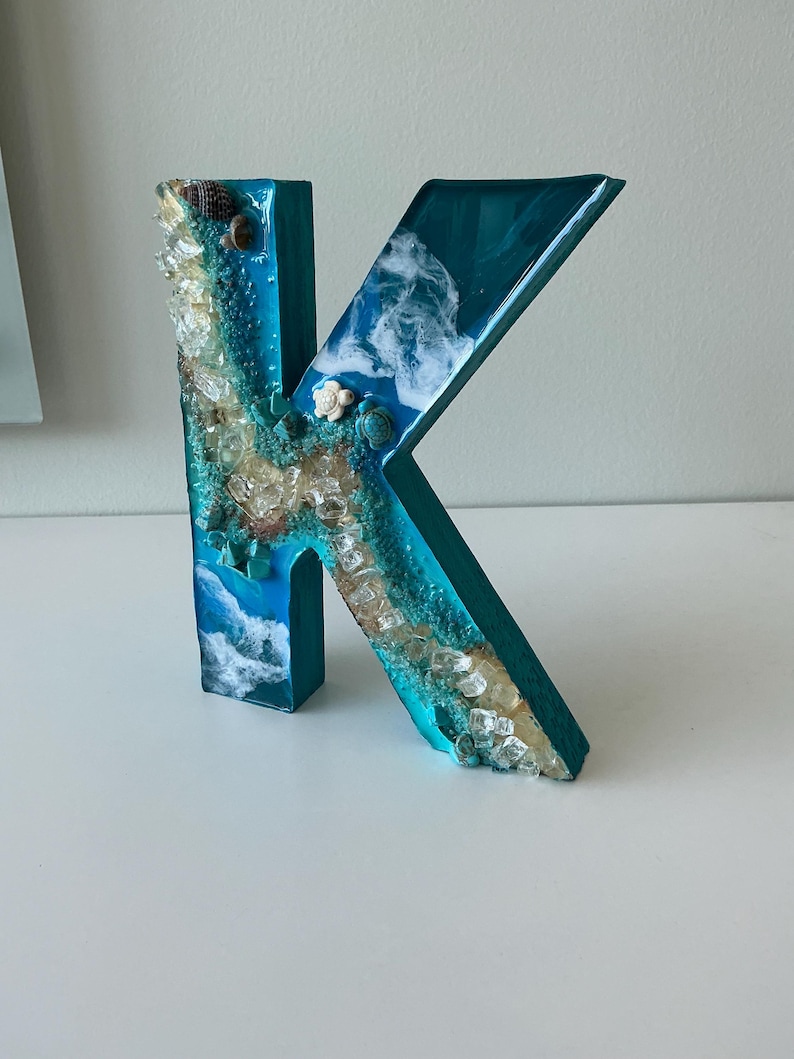 Może przedstawiać: Turkusowa drewniana litera "K" ozdobiona żywicą i elementami morskimi. Litera ma turkusową podstawę z żywicy z zatopionymi muszlami, przezroczystymi kryształami i białymi wzorami fal, tworzącymi estetykę nadmorską. Litera ma około 20 cm wysokości.