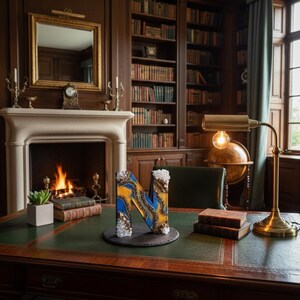 Puede incluir: Una letra decorativa "N" hecha de resina, con remolinos azules y amarillos, e incrustada con cristales. La letra est&aacute; sobre un posavasos de pizarra negra en un escritorio de cuero verde en un entorno de biblioteca. Una chimenea y estanter&iacute;as est&aacute;n en el fondo.