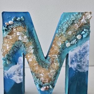 Może przedstawiać: Dekoracyjna litera "M" z motywem plażowym. Litera wypełniona jest niebieską żywicą, piaskiem, muszlami i przezroczystymi kawałkami szkła. Krawędzie są ciemnoniebieskie, a wnętrze ma wz&oacute;r fal.