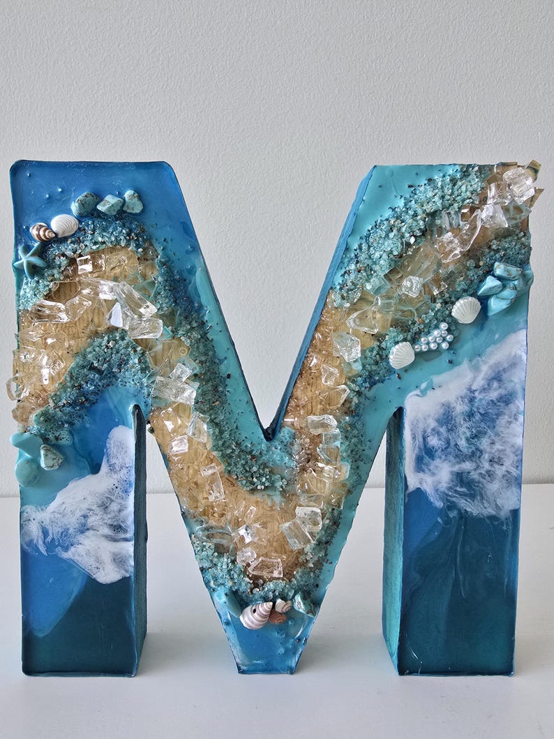 Może przedstawiać: Dekoracyjna litera "M" z motywem plażowym. Litera wypełniona jest niebieską żywicą, piaskiem, muszlami i przezroczystymi kawałkami szkła. Krawędzie są ciemnoniebieskie, a wnętrze ma wz&oacute;r fal.