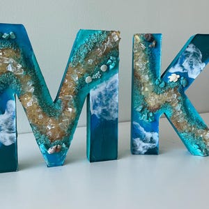 Może przedstawiać: Tr&oacute;jwymiarowe litery piszące "MK" w turkusowo-niebieskim wzorze z żywicy. Litery są ozdobione piaskiem, muszlami i przezroczystymi kryształami, co przywodzi na myśl motyw nadmorski. Litery umieszczone są na białej powierzchni.