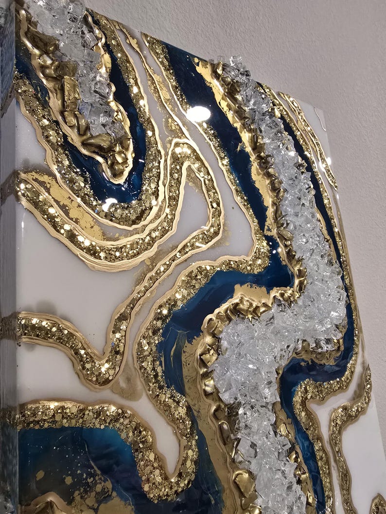 Op de afbeelding: Abstract kunstwerk van hars met een geode-ontwerp. Het kunstwerk heeft lagen van wit, diepblauw en goudkleurig glitterhars, met heldere kristalaccenten. Het stuk heeft een glanzende afwerking en een getextureerd uiterlijk.