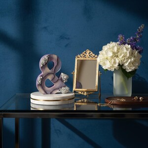 May include: A decorative ampersand sculpture in shades of purple and white, adorned with silver accents, rests on a marble base. A gold-framed mirror, a vase of white hydrangeas, and jewelry are also displayed on a glass table against a blue backdrop.