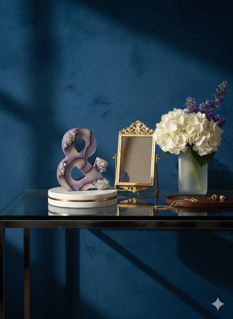 May include: A decorative ampersand sculpture in shades of purple and white, adorned with silver accents, rests on a marble base. A gold-framed mirror, a vase of white hydrangeas, and jewelry are also displayed on a glass table against a blue backdrop.