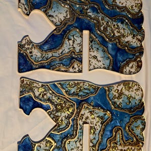 May include: Three-piece set of decorative resin art. The pieces feature a blue, white, and gold design with a glittery, layered effect. The edges are outlined in gold, creating a unique, artistic look. The pieces are designed to stand upright.