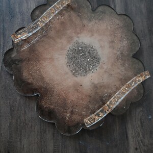 K&ouml;nnte beinhalten: Ein dekoratives Tablett aus Kunstharz mit gewelltem Rand. Das Tablett hat eine braune und beige Farbgebung mit Goldblatt-Akzenten in den Griffen. Die Mitte des Tabletts ist mit silbernem Glitzer gef&uuml;llt.