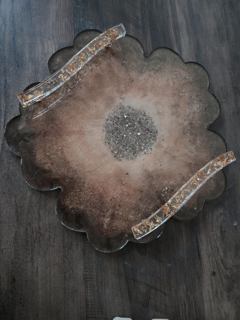 K&ouml;nnte beinhalten: Ein dekoratives Tablett aus Kunstharz mit gewelltem Rand. Das Tablett hat eine braune und beige Farbgebung mit Goldblatt-Akzenten in den Griffen. Die Mitte des Tabletts ist mit silbernem Glitzer gef&uuml;llt.